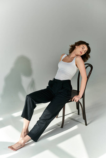 Μια νεαρή γυναίκα αποπνέει κομψότητα καθώς κάθεται σε μια καρέκλα, επιδεικνύοντας μια χαριτωμένη πόζα σε ένα σκηνικό στούντιο.. - Φωτογραφία, εικόνα