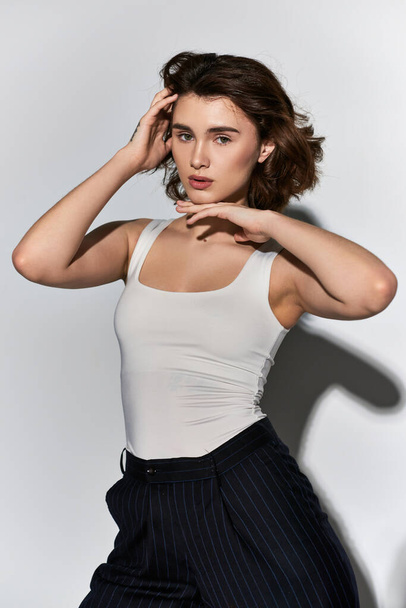 A pretty young woman posing confidently in a white tank top and black pants in a studio setting on a grey background. - Photo, Image