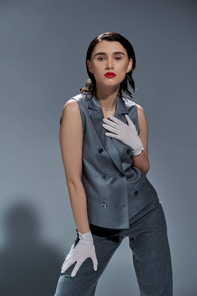 Stylish young woman strikes a pose in an elegant gray suit with vest and white gloves on a grey studio background. - Photo, Image