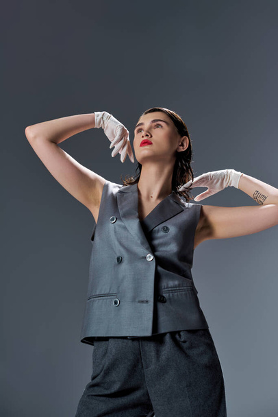 Stylish young woman strikes a pose in an elegant black suit with vest and white gloves, set in a studio against a grey background. - Photo, Image