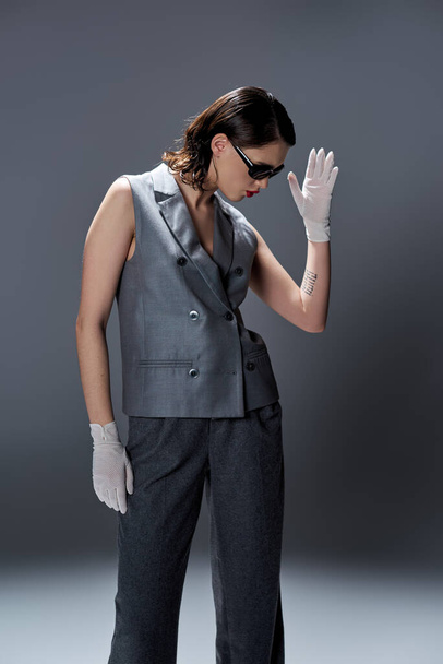 Stylowa młoda kobieta elegancko pozuje w szarym garniturze z kamizelką i białymi rękawiczkami w studio na szarym tle. - Zdjęcie, obraz