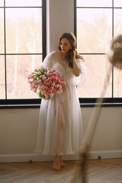 Portrait de femme enceinte en robe blanche transparente avec bouquet de tulipes roses posant près de la fenêtre dans le studio. Joyeux concept de maternité. Saison de printemps - Photo, image