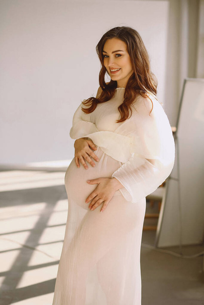 Vertical portrait of happy pregnant woman in stylish ivory dress in the studio. Beauty and fashion. Happy motherhood concept. Copy space for text - Photo, Image