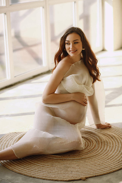 Allegro donna incinta in elegante abito avorio seduto sul pavimento e in posa in studio. Copia spazio per testo. Felice concetto di maternità - Foto, immagini