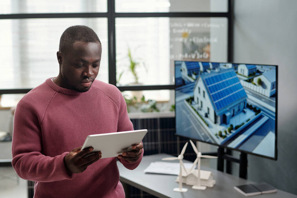 Afroamerikanische männliche Mitarbeiter in Freizeitkleidung vernetzen sich oder scrollen mit interaktiver Tafel durch Projektpunkte gegen ihren Arbeitsplatz - Foto, Bild
