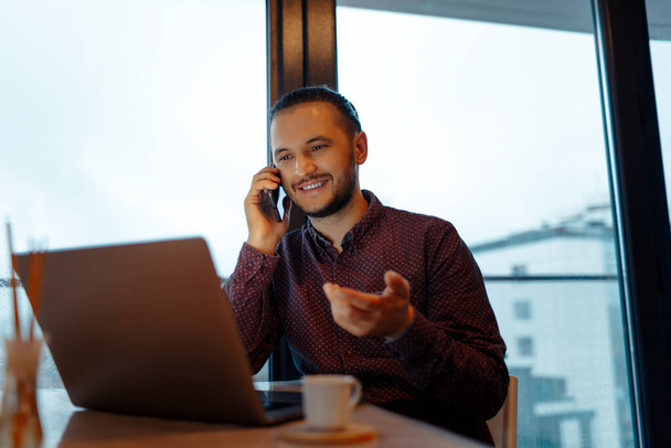 Portrait of smiling handsome man working at laptop, talking on smartphone, on background of panoramic window. - Photo, Image