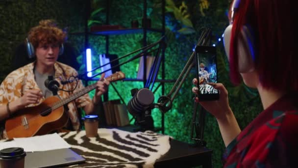Fiatal blogger a fejhallgatóban gitározik és mikrofonban énekel a stúdióban, miközben a barátnője telefonnal filmezi a közösségi médiának. - Felvétel, videó