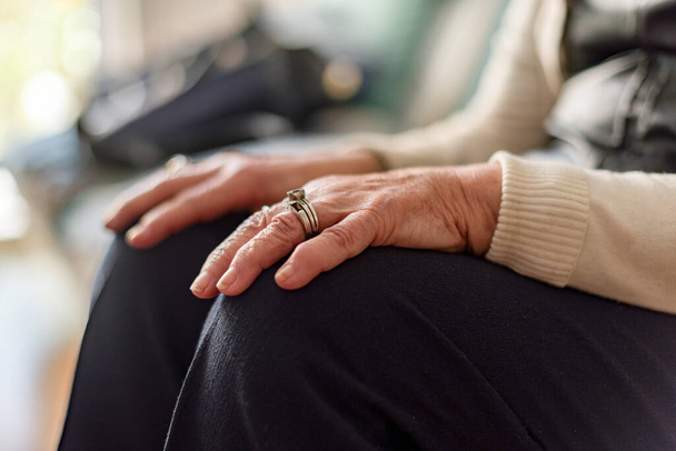 An elderly woman sits on a couch with her hands resting on her knees, wearing a ring on her left hand. Loneliness and old edge concept - Photo, Image