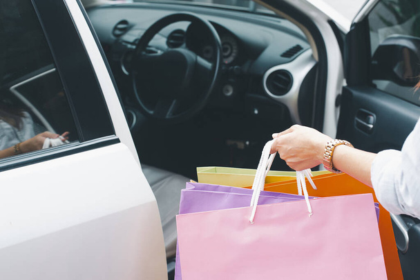 Ένα κοντινό πλάνο με το χέρι μιας γυναίκας να κρατάει πολύχρωμες τσάντες δίπλα σε ένα αυτοκίνητο, συμβολίζοντας ένα επιτυχημένο ταξίδι για ψώνια. - Φωτογραφία, εικόνα