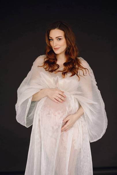 Ελκυστική έγκυος με ιβουάρ φόρεμα που ποζάρει στο στούντιο σε μαύρο φόντο. Η έννοια της ευτυχισμένης μητρότητας. Κομψό έγκυος γυναίκα  - Φωτογραφία, εικόνα