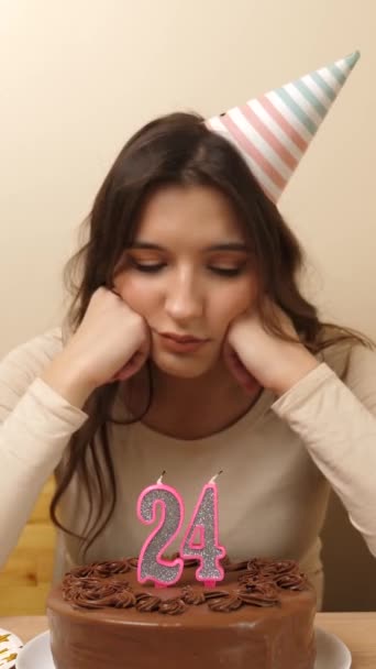 Una chica molesta se sienta frente a una mesa con un pastel festivo, en el que una vela en forma de número 24 está atascada. El concepto de una celebración de cumpleaños. vídeo vertical - Metraje, vídeo