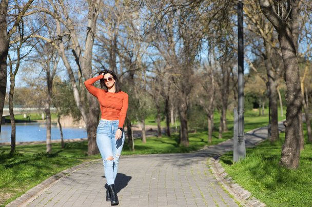 Mladá a krásná brunetka žena oblečená v neformálním oblečení a slunečních brýlích, kráčí po cestě mezi opadavými stromy na podzim, v pozadí jezero - Fotografie, Obrázek