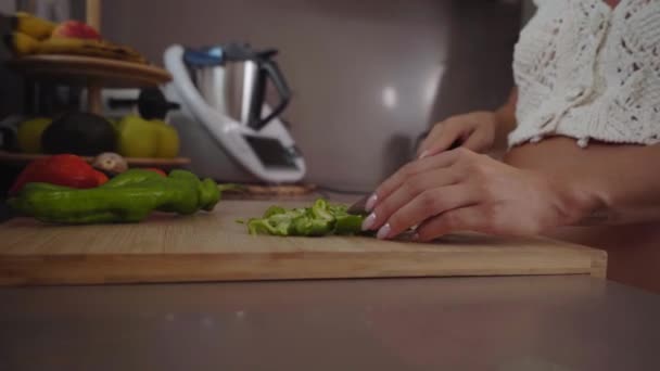 Közelkép a fiatal terhes fehér lányról, aki otthon késsel vagdossa a zöldségeket a deszkán. Az emberek főznek egy lakás konyhájában. A lány vegetáriánus ételt készít. Házimunka. - Felvétel, videó