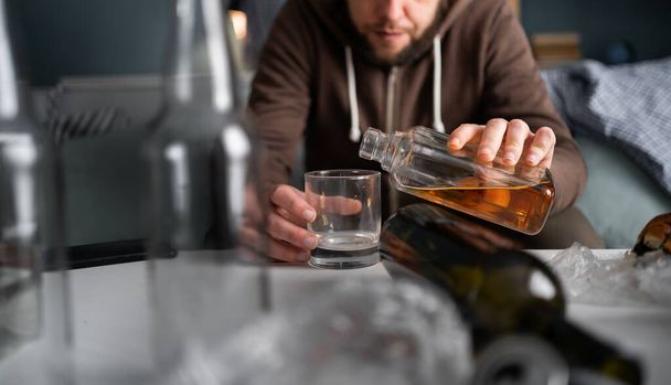 Lähikuva laukaus alkoholisti kaatamalla viskiä lasiin pöydälle istuessaan pullojen ja likaa olohuoneessa. Alkoholiriippuvuuden ja psykologisten ongelmien käsite - Valokuva, kuva