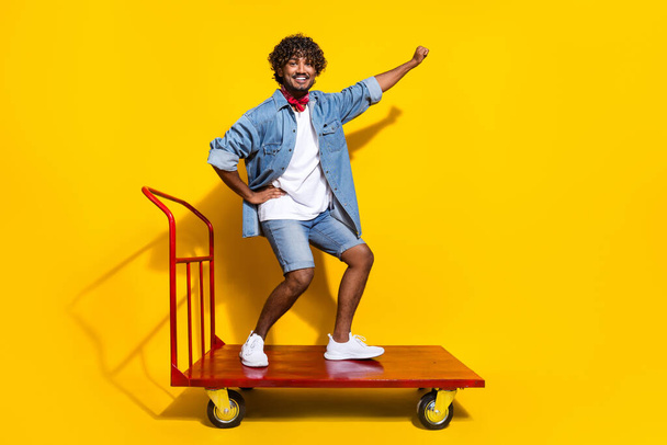 Full length photo of handsome young guy riding cart warehouse wear trendy denim garment red scarf isolated on yellow color background. - Photo, image