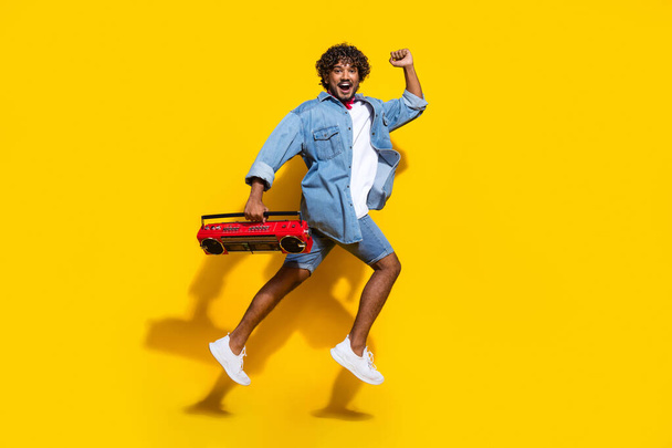 Full length photo of handsome young guy running hold boombox dressed stylish jeans garment red scarf isolated on yellow color background. - Photo, image