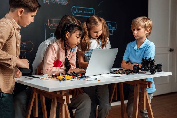 Skilled kids learning to program remote car. Schoolgirl in pink cloth use laptop for coding. Other child watch her code while teacher watch her. motherboard and electric wire also on table. Erudition. - Photo, Image
