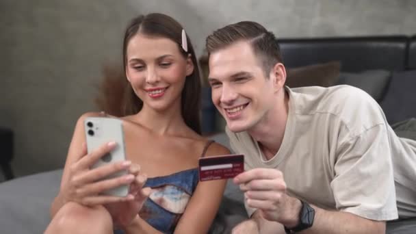 Молодая пара сидит в своей спальне, используя онлайн-приложение для оплаты и цифровой кошелек на смартфоне, чтобы заплатить кредитной картой. Электронная коммерция и современные покупки через мобильный интернет. Adit - Кадры, видео