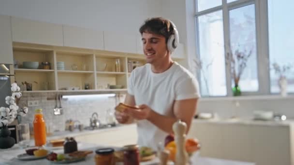 近代的なアパートのキッチンでヘッドフォンで料理を踊っているおかしい男. ワイヤレスイヤホンの朝食リスニング音楽のためのトーストを準備する陽気なリラックスした男. ハッピーハンサム 楽しむ 歌. - 映像、動画