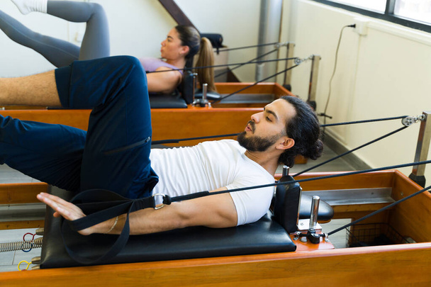 Молодой спортсмен выполняет упражнения на пилатесе реформаторской кровати в яркой тренажерной зале - Фото, изображение