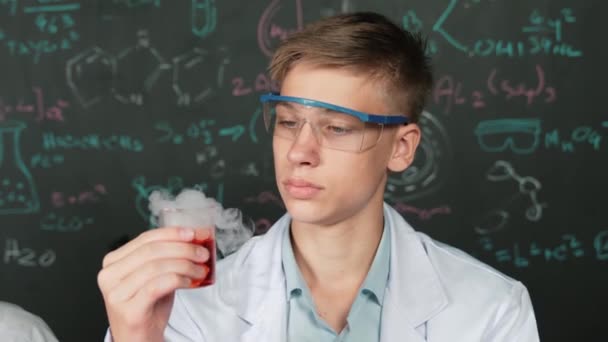 Closeup van jongen inspecteren chemische oplossing terwijl het houden van beker op blackboard met chemische theorie. Slimme wetenschapper doet experiment terwijl analyse gekleurde oplossing terwijl het dragen van laboratoriumjas. Edificatie - Video