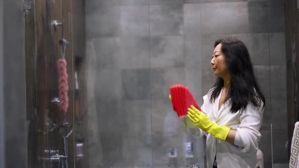 Een meisje veegt en stoffeert douchecabine in de badkamer. Schoonmaakconcept. Huisvrouw werk Jonge Aziatische vrouw reinigt glas in de badkamer. De meid maakt het huis schoon.. - Video