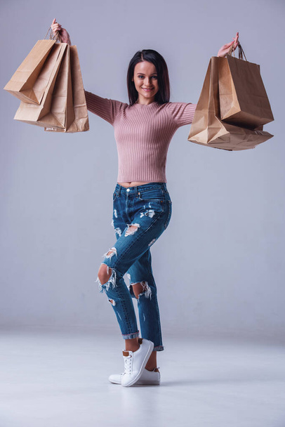 Полный портрет привлекательной молодой женщины в повседневной одежде, держащей сумки с покупками, смотрящей в камеру и улыбающейся, на сером фоне
 - Фото, изображение