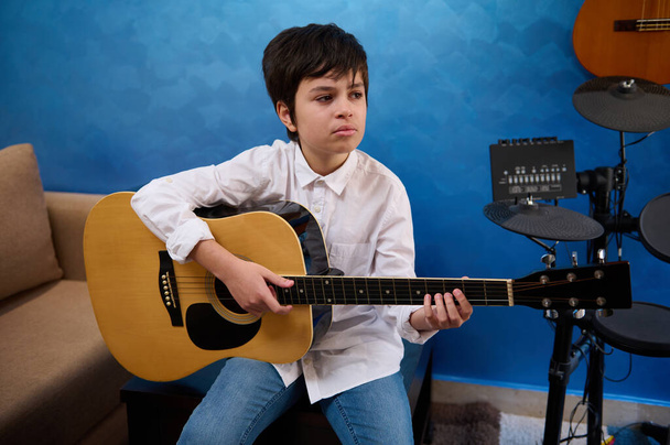 Teenager boy playing acoustic guitar at home, sitting against musical instruments and blue wall background. Child learning classical chord instrument. People. Education. Music. Hobbies and leisure - 写真・画像