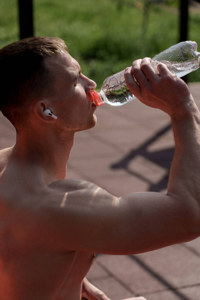 Un homme en forme et athlétique prend une boisson rafraîchissante dans une bouteille d'eau claire, illustrant la santé et la forme physique lors d'une séance d'entraînement en plein air. - Photo, image