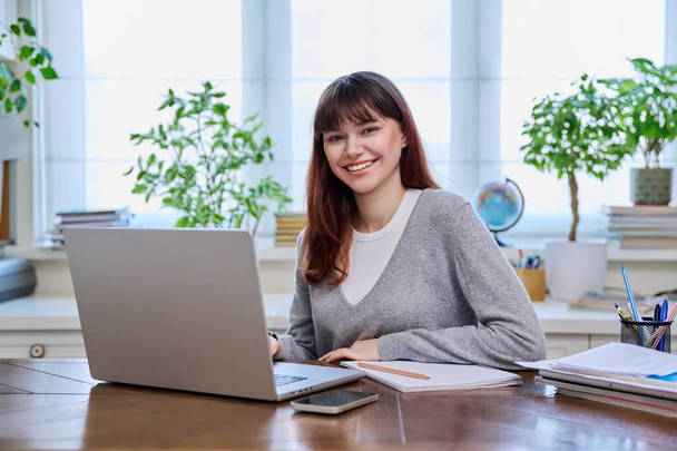 Young female college student studying at home at desk using computer laptop, writing in notebook, smiling looking at camera. E-learning, education, technology, knowledge, youth concept - Фото, зображення