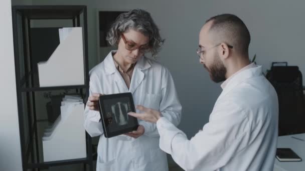 Střední záběry zralého bělošského lékaře v bílém plášti a kolegy z Blízkého východu stojícího společně v kanceláři, držícího tablet s rentgenovým obrazem, diskutujícího o vzácném lékařském případu - Záběry, video