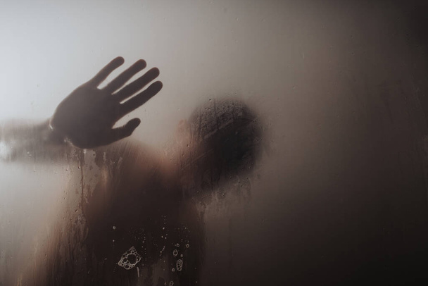 Mystérieuse silhouette d'une personne vue à travers une surface en verre embrumé avec une empreinte claire de la main. - Photo, image