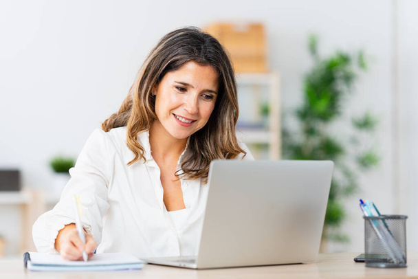 Mujer rubia adulta tomando notas mientras trabaja desde casa - Foto, imagen