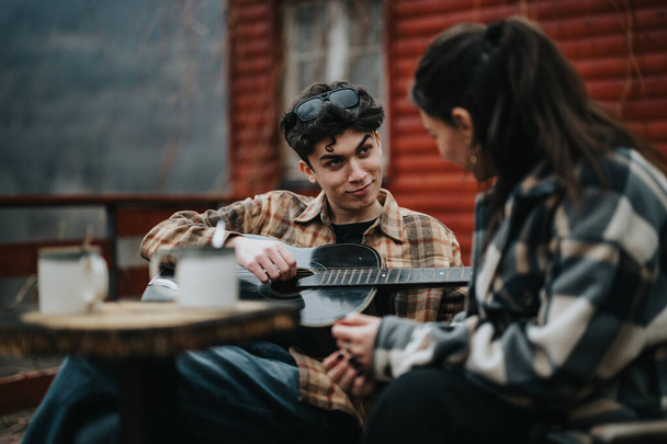 Young couple sharing a moment over a guitar song, enjoying warm drinks on a cozy terrace. - Photo, Image