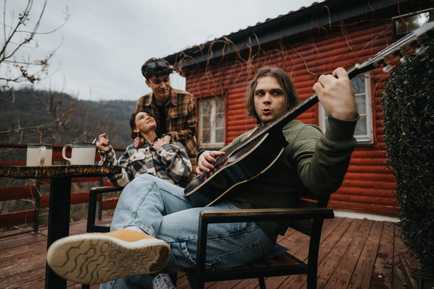 Les jeunes adultes se détendent avec des guitares et du café sur un porche en bois près d'une cabane rustique, partageant un moment de musique et de rire. - Photo, image