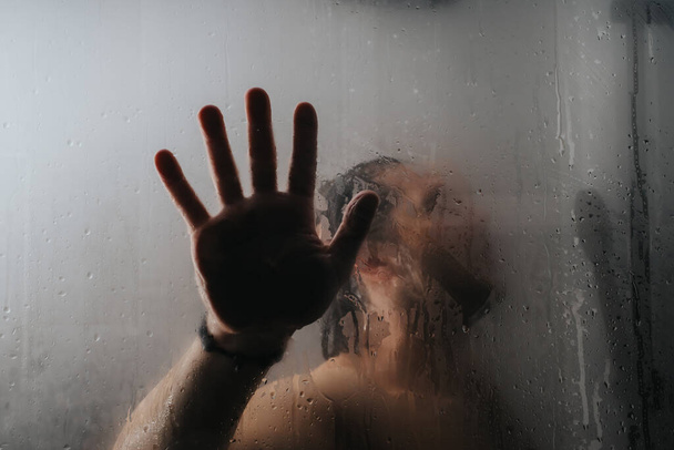 Silueta de una persona contemplativa vista a través de una niebla, puerta de ducha de vidrio húmedo, con una mano presionada contra la superficie. - Foto, imagen