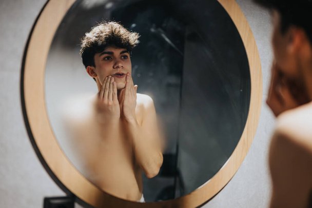 Gömleksiz genç erkek dairesel aynada yansımasına bakıyor, modern bir banyo ortamında yüzüne dokunuyor.. - Fotoğraf, Görsel