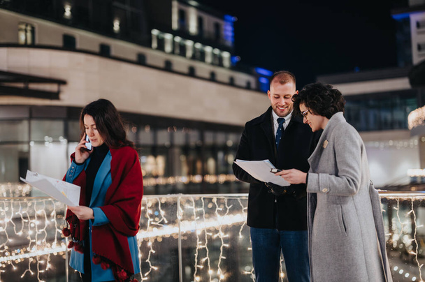 Üç profesyonel kış boyunca dışarıda toplandı ve arka planda festival ışıkları olan gazeteleri tartıştılar.. - Fotoğraf, Görsel