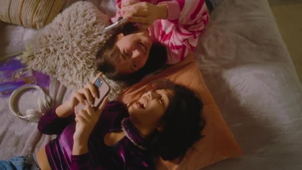 Zwei multiethnische beste Freunde im Schlafzimmer lachen und scrollen mit dem Handy durch die sozialen Medien. Glückliche Teenager-Mädchen verbringen ihre Freizeit zu Hause auf dem Bett liegend. Bestien haben gemeinsam Spaß. - Filmmaterial, Video