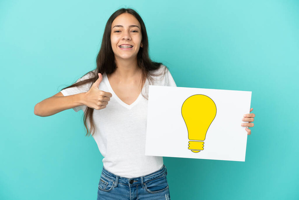 Young French woman isolated on blue background holding a placard with bulb icon with thumb up - Photo, Image