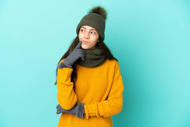 Young French girl isolated on blue background with winter hat having doubts while looking up - Photo, Image