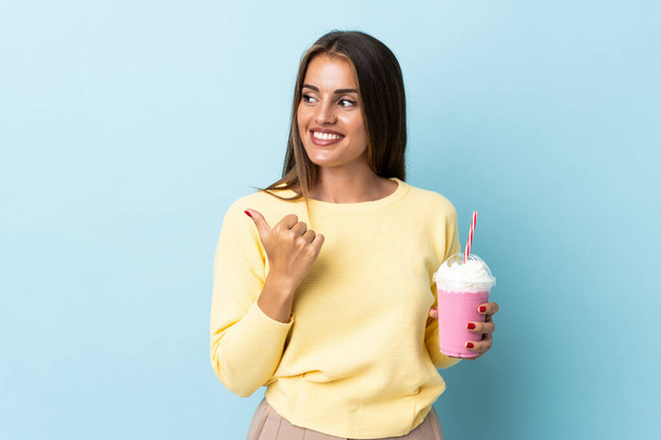 Jonge Uruguayaanse vrouw met aardbeienmilkshake geïsoleerd op blauwe achtergrond wijzend naar de zijkant om een product te presenteren - Foto, afbeelding