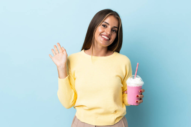 Jonge Uruguayaanse vrouw met aardbeien milkshake geïsoleerd op blauwe achtergrond salueren met de hand met vrolijke uitdrukking - Foto, afbeelding