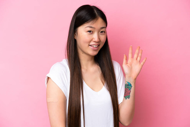 Pembe arka planda yara bandı takan genç Çinli kadın mutlu bir ifadeyle selam veriyor. - Fotoğraf, Görsel