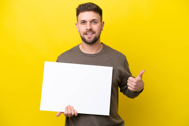 Νεαρός καυκάσιος άνδρας απομονωμένος σε κίτρινο φόντο κρατώντας ένα άδειο πλακάτ με τον αντίχειρα σηκωμένο - Φωτογραφία, εικόνα