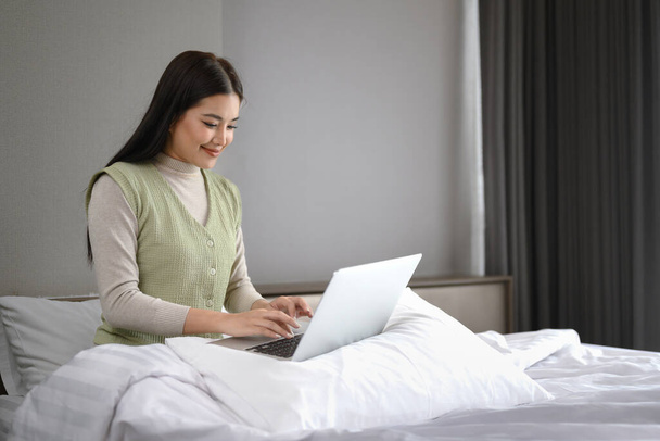Krásná asijská žena v ležérní oblečení pracuje s notebookem sedí na posteli doma. - Fotografie, Obrázek