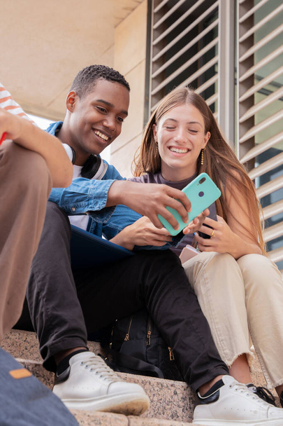 Uśmiechnięci wielorasowi studenci siedzą na zewnątrz i rozmawiają przez telefon komórkowy. Koncepcja życia uniwersytetu - Zdjęcie, obraz