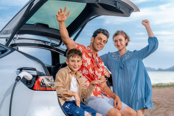 Rodinná dovolená cestování na pláži s elektrickým autem, krásná rodina sedí na kufru, nabíjení EV autobaterie se zelenou a udržitelnou energií. Rodinné cestování a ekologicky šetrné auto. Perpetual - Fotografie, Obrázek