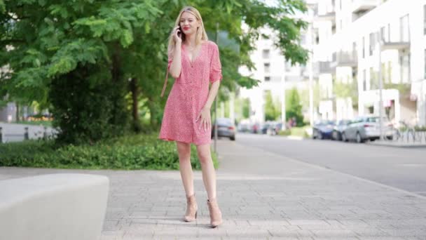 Radosna młoda kobieta w różowej letniej sukience rozmawiająca przez telefon, stojąca na miejskim chodniku z drzewami i budynkami w tle. - Materiał filmowy, wideo