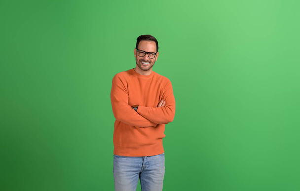 Młody przystojny przedsiębiorca stojący z skrzyżowanymi ramionami i uśmiechnięty do kamery na zielonym tle - Zdjęcie, obraz
